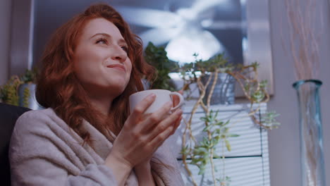 a young caucasian woman is sitting at home cozy with a blanket, sipping coffee or tea, happy