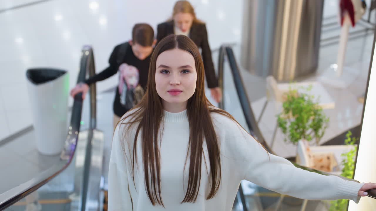 Elegant woman in white sweater ascends escalator in modern shopping mall, she gazes ahead while two people stand behind her. Bright indoor lighting, sleek design, and relaxed ambiance