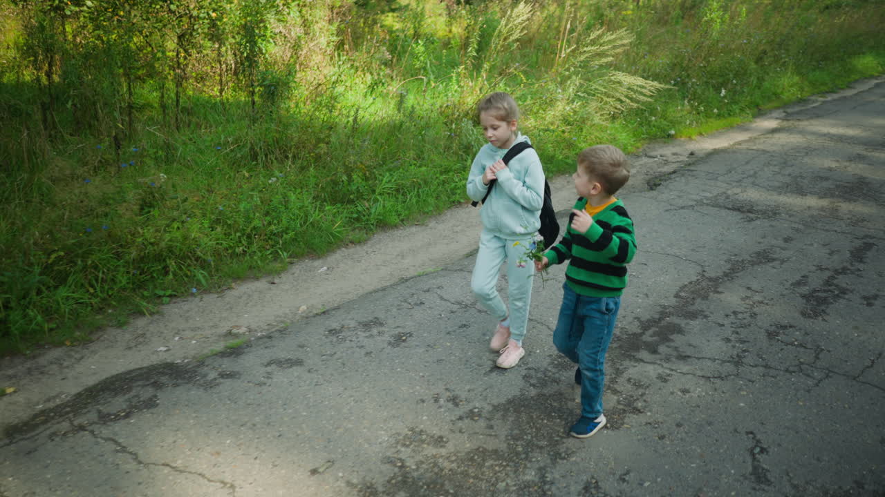 Little girl in light tracksuit adjusts bag strap while walking beside younger brother holding wildflowers along quiet cracked road bordered by tall grass and green forest under soft daylight