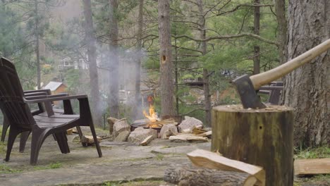 an axe is embedded in a log near a blazing fire pit, surrounded by chopped wood and plastic chairs, set in a forested area