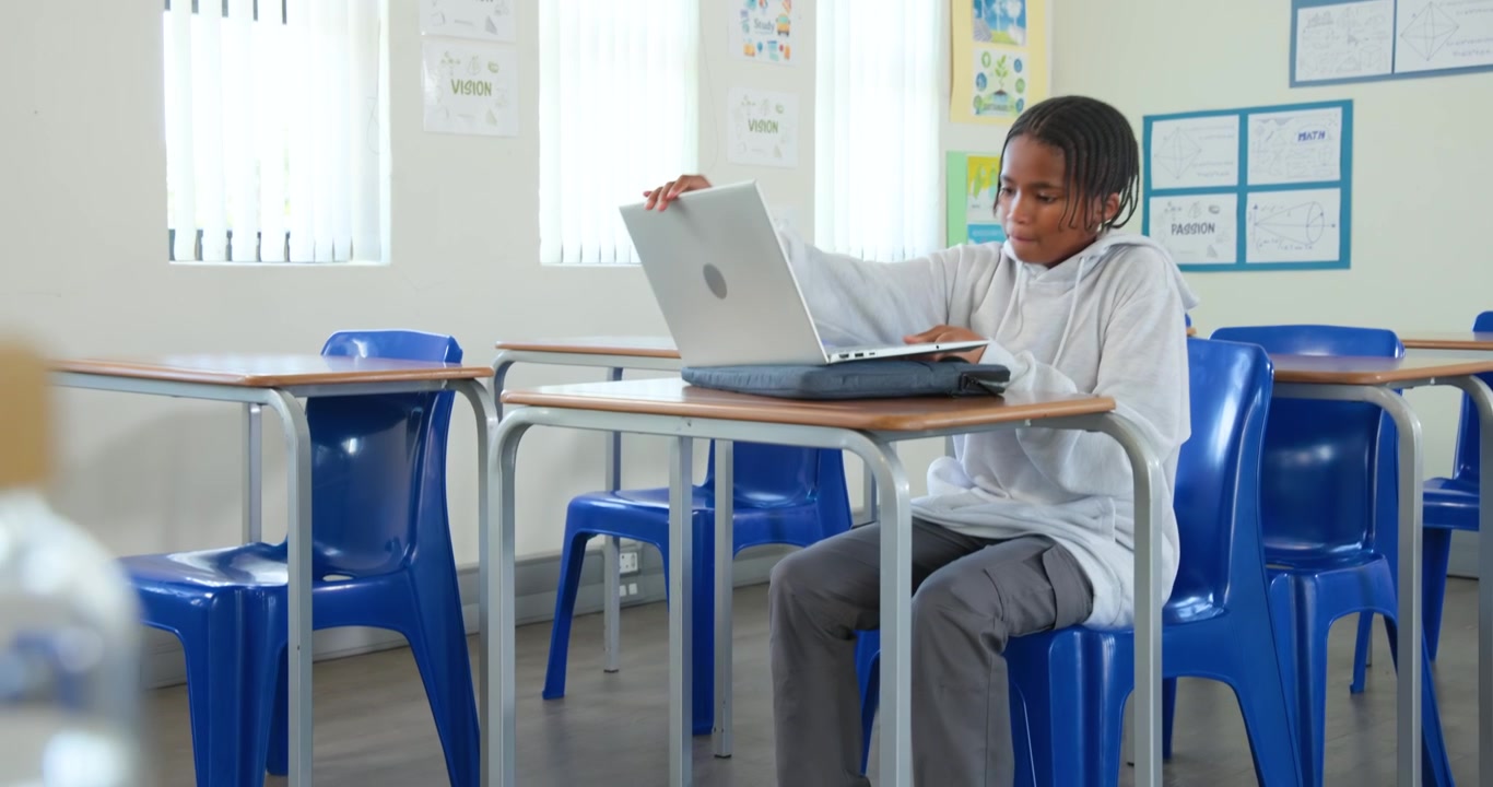 Adolescente africano che entra in fondo all'aula, posa lo zaino, tira una sedia, apre il laptop e si prepara a studiare