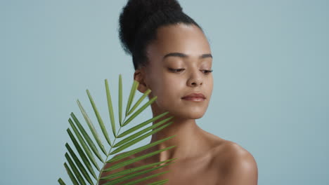 retrato de una hermosa mujer afroamericana tocando la cara con una hoja acariciando una piel suave y saludable disfrutando de una belleza natural juguetona sobre un fondo azul