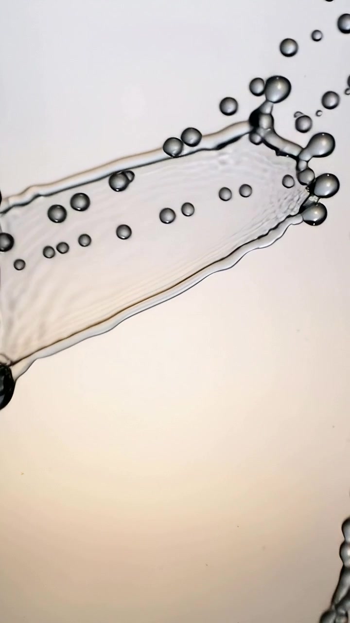 Video verticale: Foglio di liquido trasparente ad arco che scorre da un ugello sinistro in studio, formando goccioline.