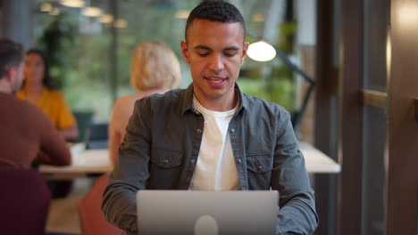 Young-Businessman-Working-On-Laptop-At-Desk-In-Office-Pausing-To-Look-Out-Of-Window