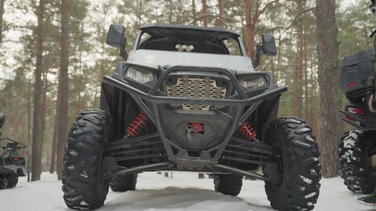 Primo piano della parte anteriore di un UTV in una foresta innevata, griglia meccanica e paraurti rinforzato visibili, i piloti in tenuta mimetica e caschi si consultano, dettagli del verricello e delle sospensioni in mostra, spedizione di gruppo che si prepara per il fuoristrada.