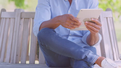 close up of man sitting on bench under tree in summer park using digital tablet