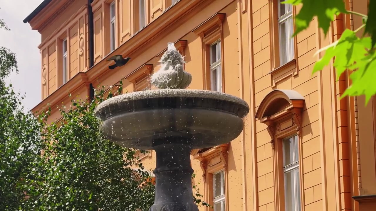 Fuente de piedra salpicando frente a una cálida fachada histórica naranja en el casco antiguo de Liubliana, con hojas verdes y ambiente de ciudad veraniego.