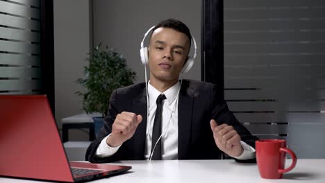 young successful african businessman in suit listening to music on the computer in headphones, dancing in the office. 60 fps