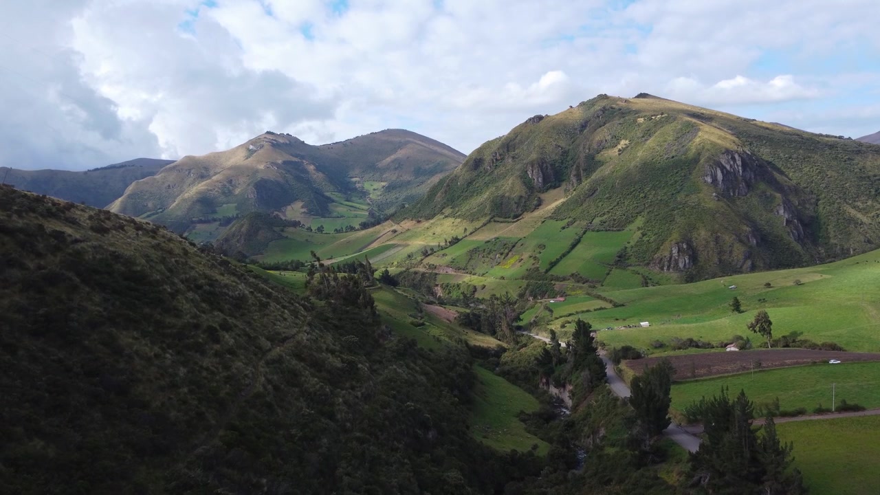 Veduta aerea con drone di una valle andina verde e soleggiata in Ecuador, con piccole fattorie, campi arati e montagne ondulate sotto un cielo luminoso con nuvole bianche