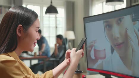 asian female digital editor using mobile phone while works in photo editing software on his personal computer in office