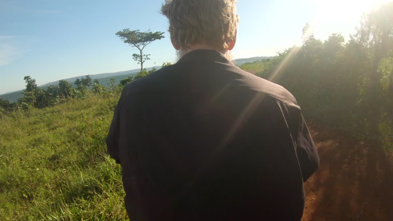 A point of view shot from behind of a ginger haired man riding a motor cycle on a rural dirt road alongside Lake Victoria in Africa.