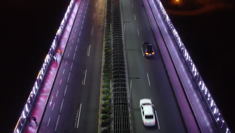 video de drones de un puente por la noche iluminado con luces moradas, la gente camina por la acera mientras los autos pasan por los carriles de la calle