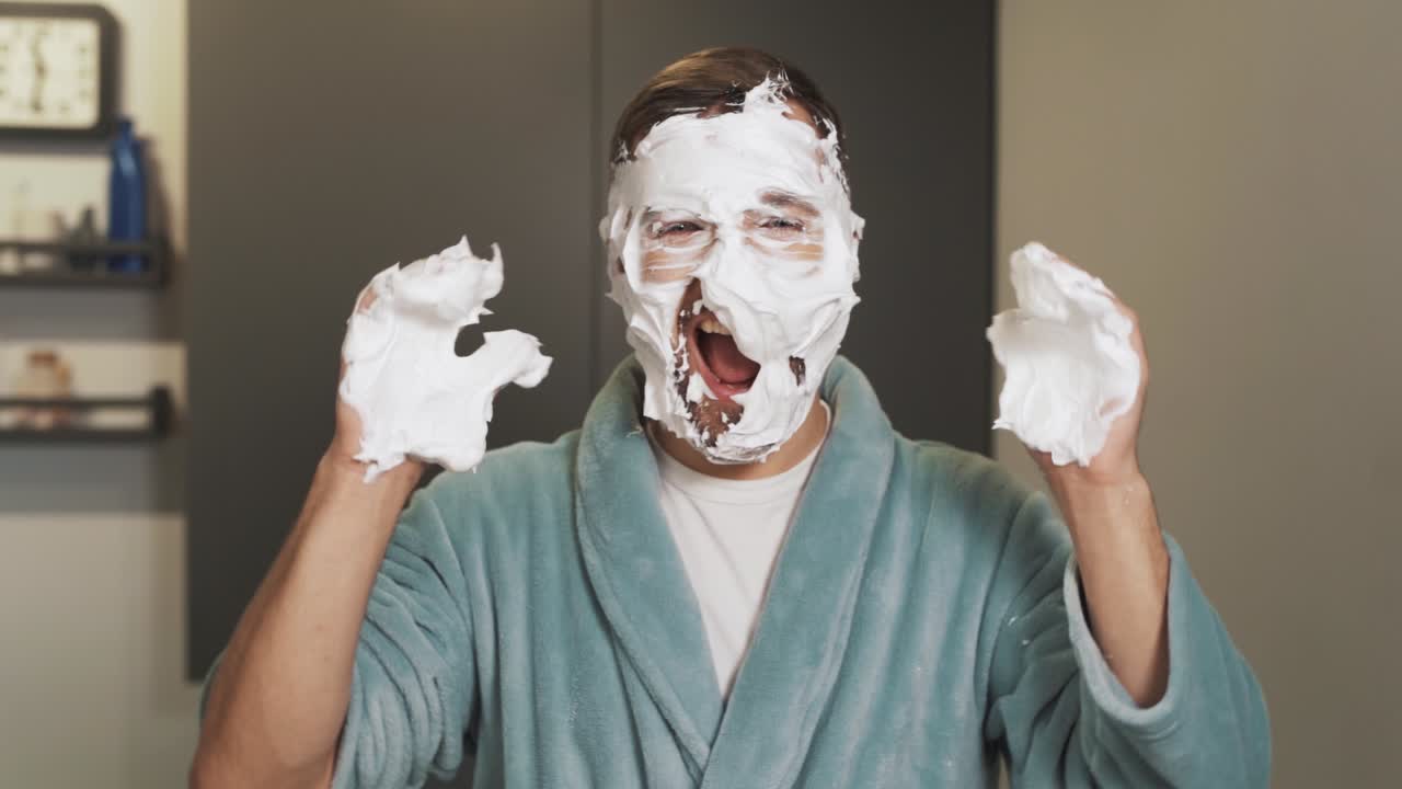 얼굴에 거품이 있는 명랑한 남자가 얼굴에 크림을 문지르면서 카메라를 보고 웃는다