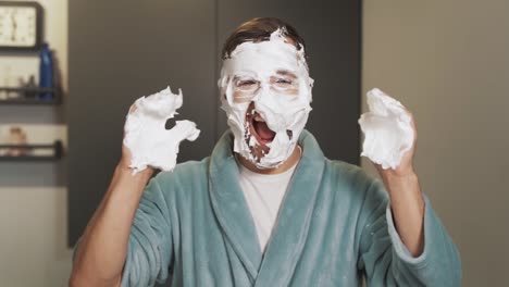 a cheerful man with foam on his face laughs at the camera while rubbing cream on his face