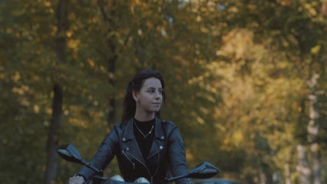 Hübsche-Lächelnde-Europäische-Junge-Frau,-Die-An-Einem-Sonnigen-Tag-Ein-Motorrad-Mit-Lederjacke-Im-Wald-Mit-Lebhaften,-Bunten-Goldenen-Herbstblättern-Fährt-4