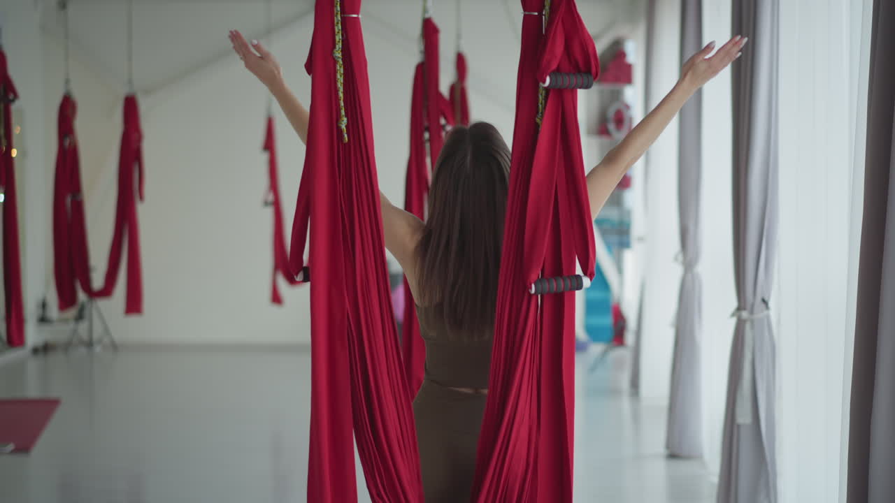 Wellness advocate lifts hands stretching while doing breathing exercise in studio among red silk hammocks focusing on balance energy mindfulness under soft natural light showing serenity and peace