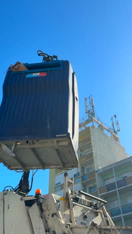 a blue waste container being emptied by a garbage truck under a clear blue sky