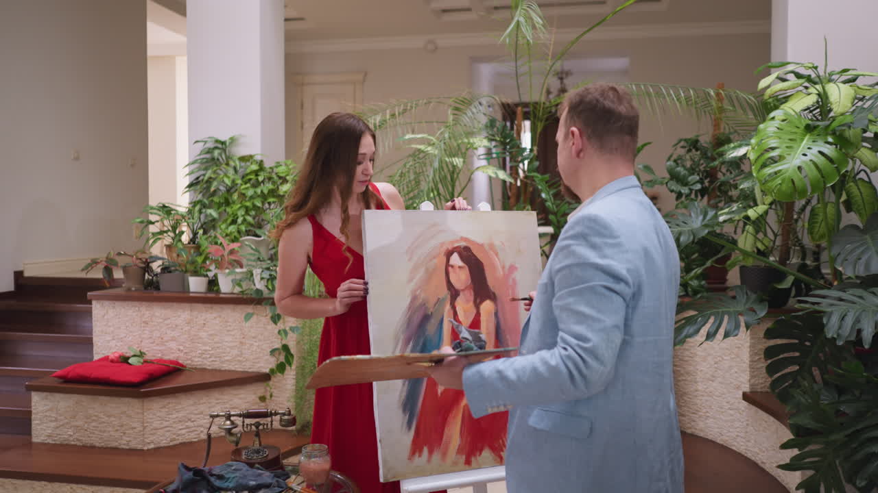 느슨한 머리카락을 가진 라이브 모델은 스에 있는 초상화를 바라보고 있습니다. 은 드레스를 입은 젊은 여성은 예술 스튜디오에서 캔버스에 나타나는 예술가의 기술과 창의성을 목격합니다.