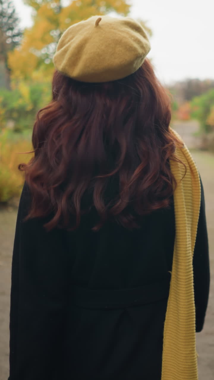 Vista trasera de una dama con una boina amarilla y una bufanda a juego, dando un paseo otoñal en un jardín, mirando a su alrededor y ajustándose su cabello rojo ondulado mientras camina.