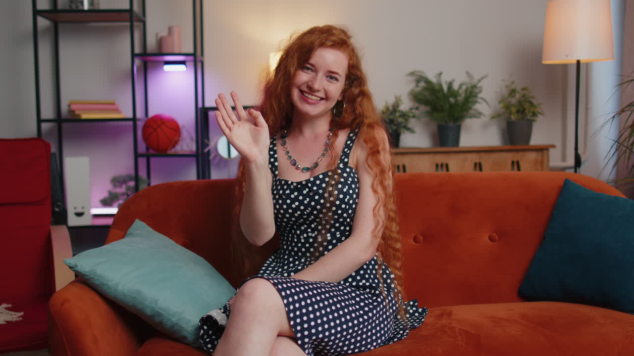 chica pelirroja sonriendo amigablemente a la cámara, agitando las manos gestando hola, hola, saludo en casa en el sofá