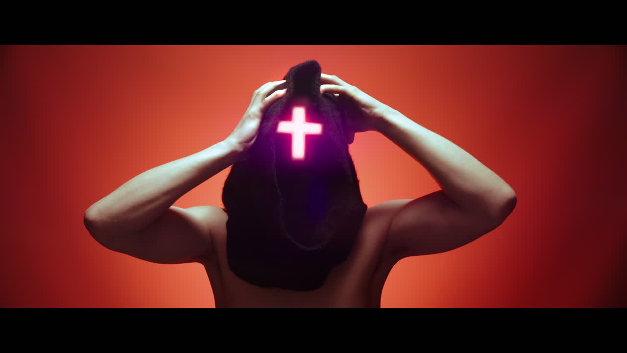 Masked man with cross of light glowing on his face grabs his head with his hand - red background