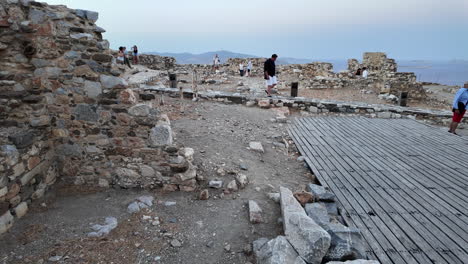 greece,astypalea, querini castle during sunset, people walking around the ruins of the castle and the white chapel with blue dome, a golden retriever is laying on the wooden floor across the chapel.