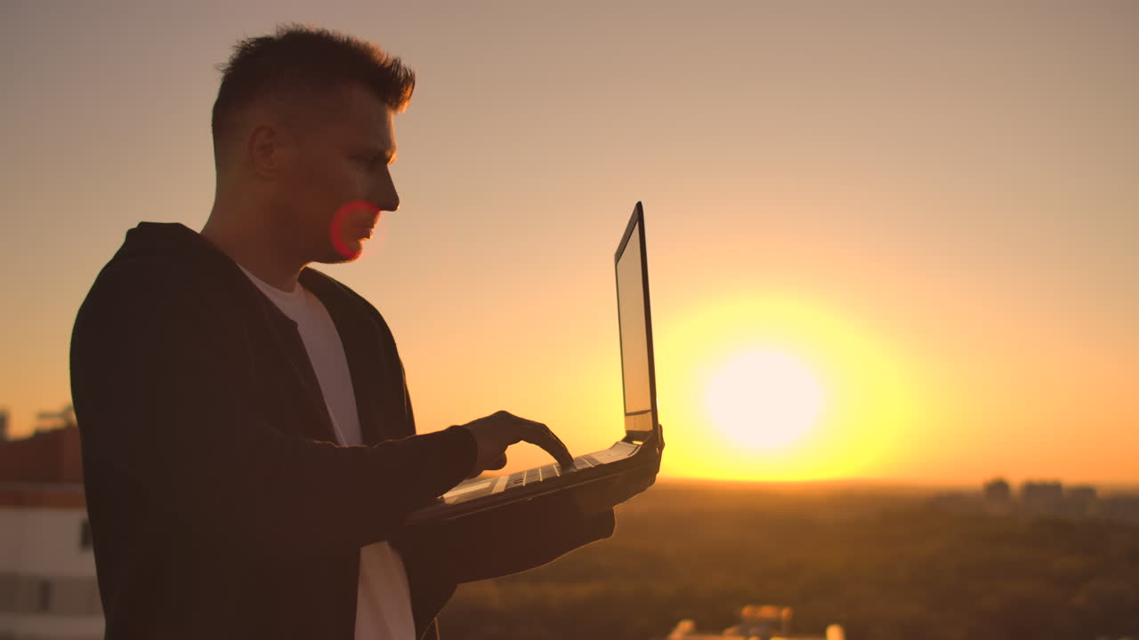 A male stockbroker freelancer stands on a rooftop at sunset with a laptop and types on a keyboard with his fingers looking at the cityscape from a bird's eye view.
