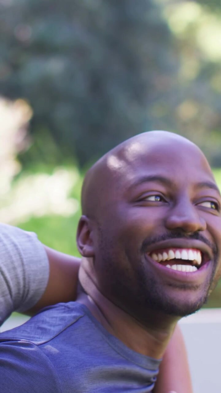 Vertical video: Laughing man in park, woman behind in gray tee wrapping arms climbing on his back