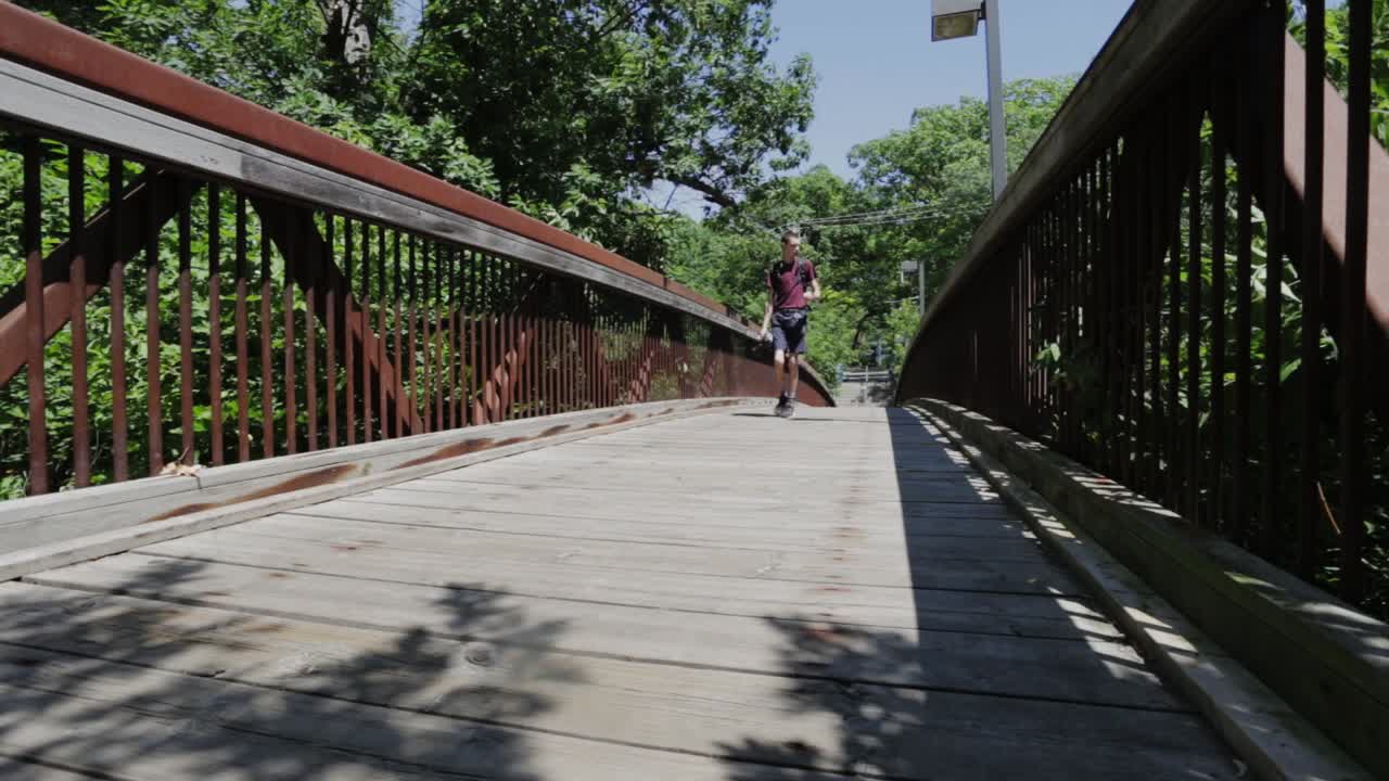 Young Man Carrying A DSLR Camera Walking On A Wooden Path - Low-Level Shot