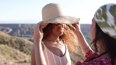 two women enjoying the outdoors, one adjusting the other's straw hat in a scenic landscape