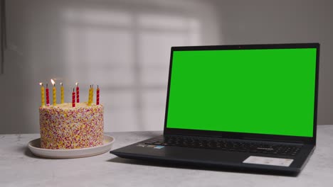 Studioaufnahme-Eines-Laptops-Mit-Grünem-Bildschirm-Neben-Einer-Geburtstagstorte-Mit-Brennenden-Kerzen,-Die-Ausgeblasen-Werden