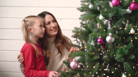 madre con su hija colgando adornos navideños en el árbol de navidad