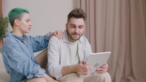 man holding a tablet while talking with his girlfriend sitting on sofa at home