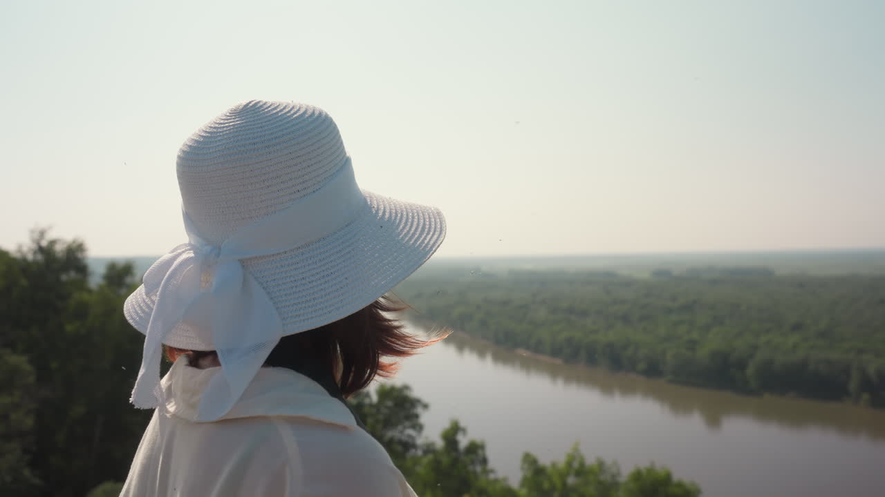 Woman wearing white sunhat and light blouse takes photo of river view from grassy field, insect flying near her hand as she stands in warm sunlight surrounded by summer greenery