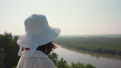 woman wearing white sunhat and light blouse takes photo of river view from grassy field, insect flying near her hand as she stands in warm sunlight surrounded by summer greenery
