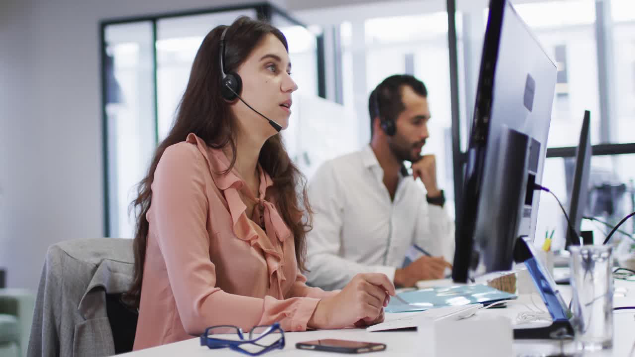 mujer de negocios caucásica sentada en el escritorio hablando usando auriculares de teléfono y computadora en una oficina ocupada