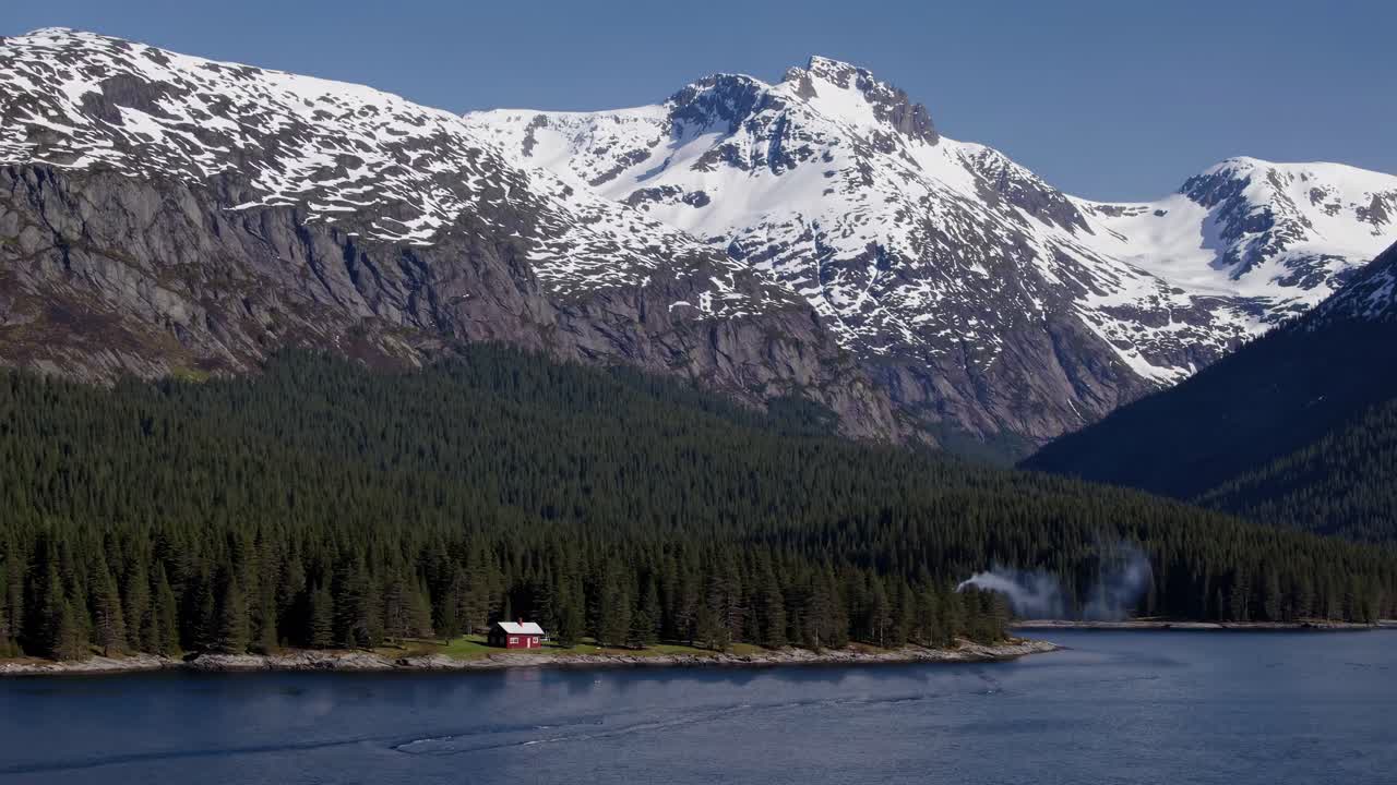 Aerial view of a serene mountain landscape with a red cabin by a lake, snow-capped peaks, and dense