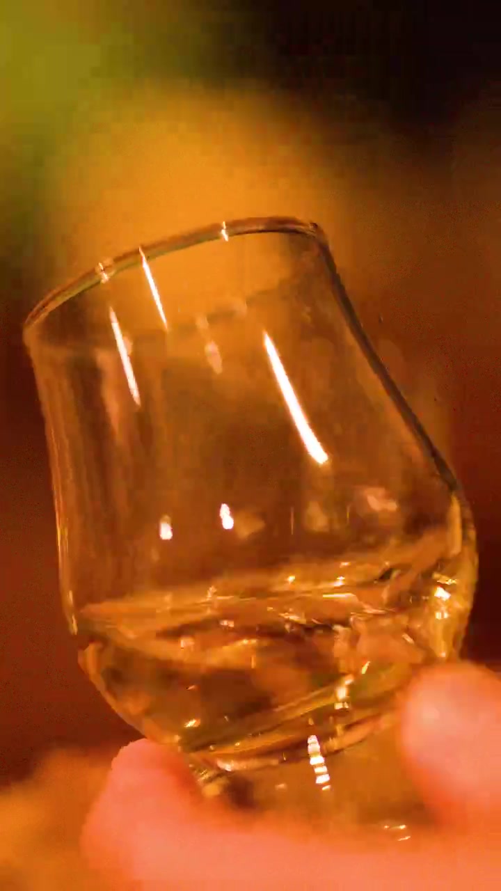 Una mano fa roteare un bicchiere di whisky, il liquido dorato cattura la calda e soffusa illuminazione del bar, con messa a fuoco ridotta.