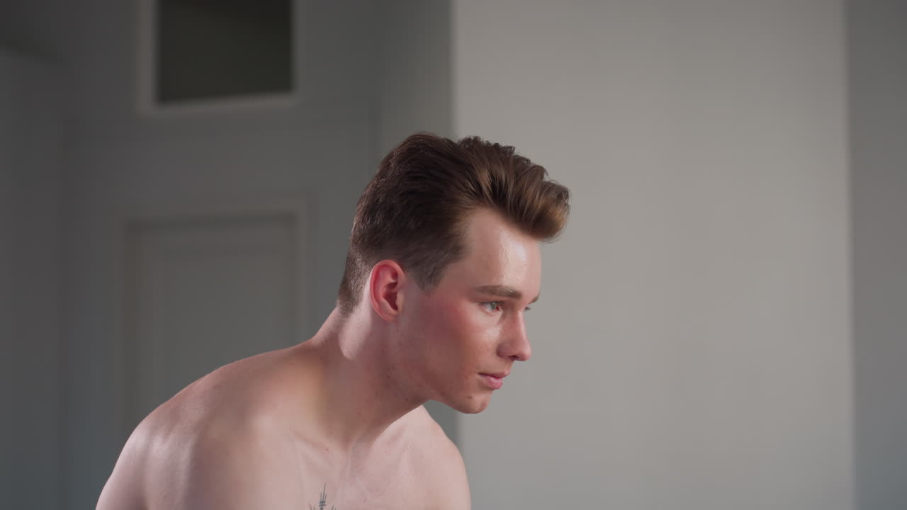 shirtless young man with slicked hair slightly bent forward turning head to look at camera in bright indoor room with plain walls and closed white door in background