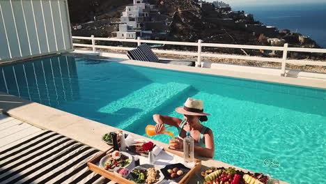 Vista-Aérea-De-Una-Chica-Feliz-En-Una-Piscina-Privada-Sirviendo-Jugo-De-Naranja-En-La-Bandeja-Del-Desayuno-En-Un-Día-Soleado-En-La-Lujosa-Villa-De-Mykonos,-A-Cámara-Lenta