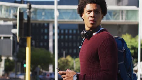 African-american-man-in-city-using-smartphone,-wearing-headphones-and-backpack-hailing-cab-in-street