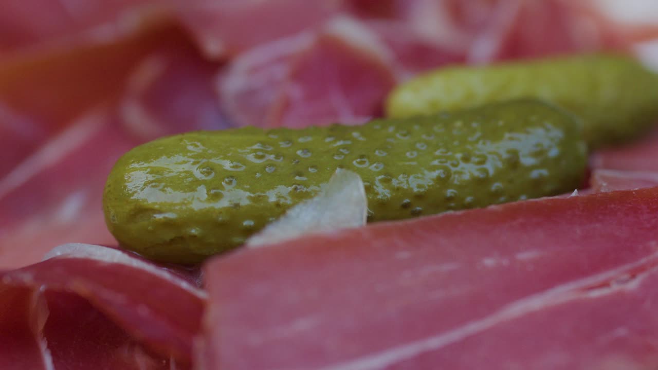 Primo piano di una mano che prende un sottaceto da un piatto di salumi con prosciutto, lattuga e verdure, luce naturale.