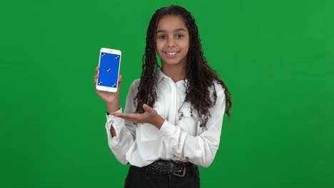 smiling positive african american teenage girl showing smartphone with blue screen posing at chromakey background. confident happy teenager advertising mobile app on green screen.