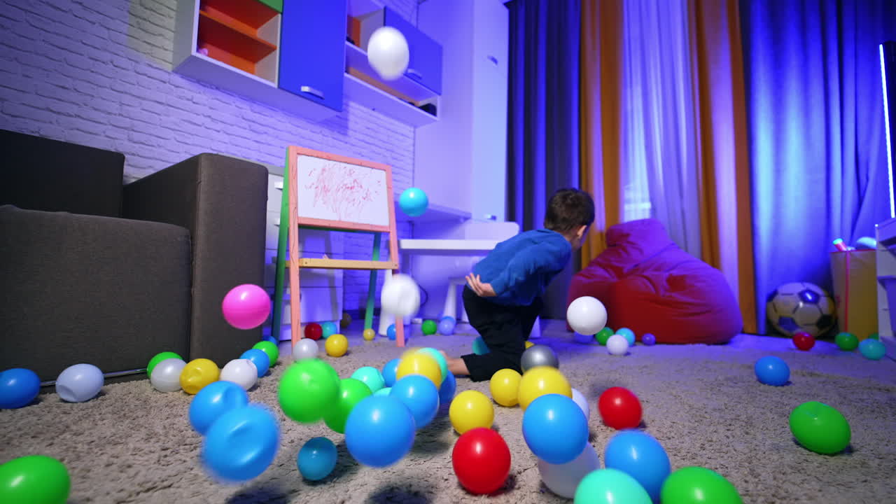 Happy kid sitting on the floor catches the balls falling on him. Baby boy crawls to the bean bag chair.