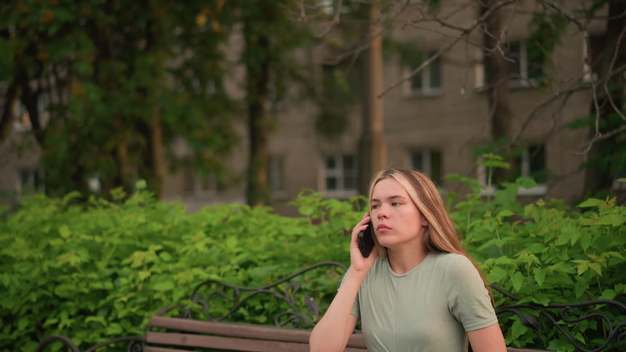 젊은 여성은 나무 벤치에 앉아 전화 통화를 하고, 머리카락을 조정하고 목에 손을 고, 울창한 초록색, 나무, 그리고 배경의 건물로 둘러싸여 있습니다.