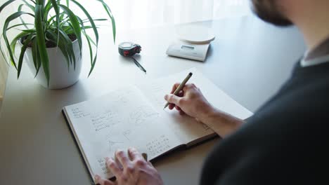 Close-up-shot-of-a-man-hand-trying-to-solve-a-geometry-problem-in-a-notebook,-with-a-indoor-plant-on-the-table-along-with-stopwatch-at-daytime