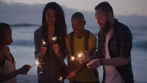 Grupo-Diverso-De-Amigos-Celebrando-La-Víspera-De-Año-Nuevo-Usando-Fuegos-Artificiales-De-Bengala-Disfrutando-Juntos-De-Una-Tranquila-Fiesta-En-La-Playa-Al-Atardecer