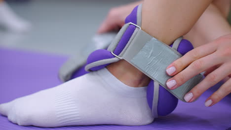 primer plano enfocado de las manos apretando una correa de peso del tobillo en un pie cubierto de calcetines, preparándose para un entrenamiento deportivo, la alfombra de ejercicio púrpura y el entorno de gimnasio interior