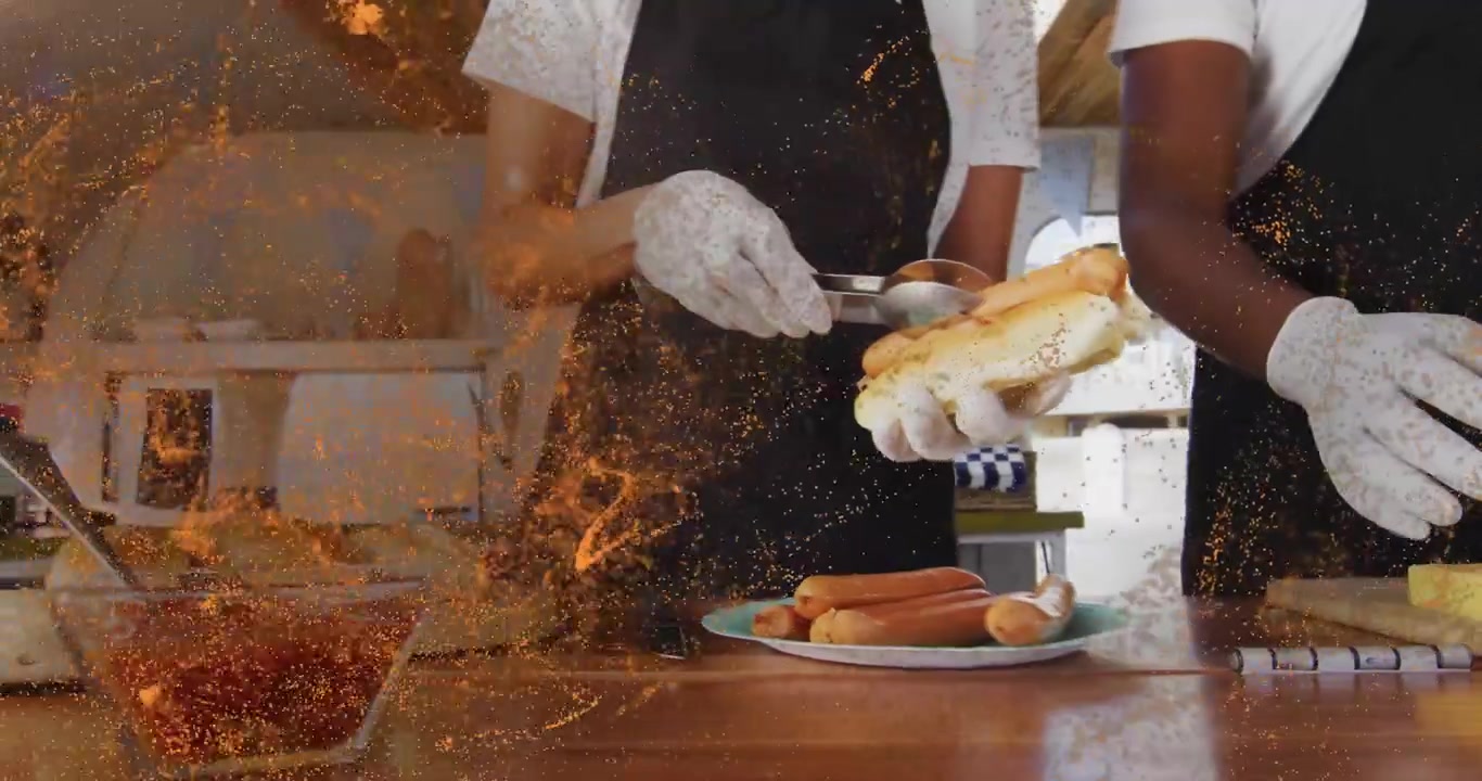 Uomo che prende un panino ai semi di papavero, mentre una donna preleva la salsa e posiziona una salsiccia con le pinze per la preparazione di hot dog.