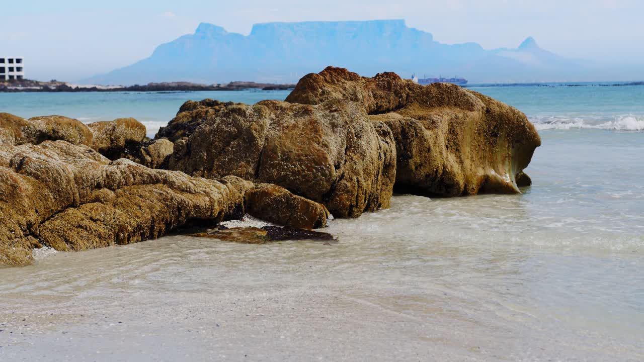 Langsamer Schwenk über Strandfelsen, Tafelberg im Hintergrund, Wellen schwappen auf den weißen Sand